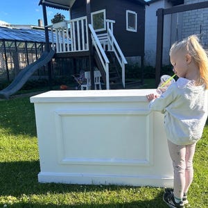 Puede incluir: Un mostrador blanco rectangular para casa de juegos con un diseño de panel enmarcado. Un niño está de pie junto al mostrador, bebiendo de un vaso transparente con una pajita. El fondo incluye una casa de juegos negra con un tobogán y un columpio.