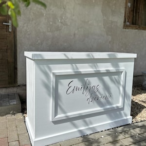 Puede incluir: Una estructura rectangular de madera blanca con un panel enmarcado. El panel presenta el texto cursivo "Emitijas dzērieni" en negro. La estructura está al aire libre, sobre una superficie de ladrillo, con una puerta y una pared al fondo.