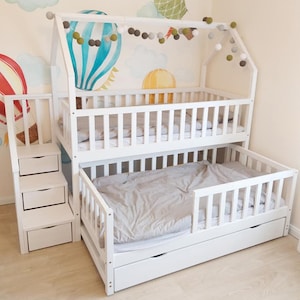 Puede incluir: Una litera de madera blanca con forma de casa, con una cama superior, una cama inferior extraíble y una escalera con cajones. La cama está colocada contra una pared con murales de globos aerostáticos y guirnaldas decorativas de bolas de fieltro.