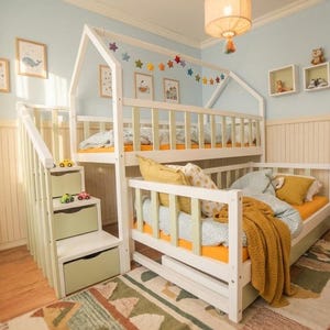 Könnte beinhalten: Ein weißes Etagenbett mit hausförmigem Rahmen, mit einem unteren Bett und einem oberen Etagenbett. Das Bett hat eine Treppe mit Schubladen und ist mit bunten Sternengirlanden und gerahmten Kunstwerken dekoriert. Der Raum hat eine hellblaue Wand.