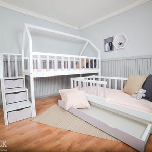 Puede incluir: Una litera de madera blanca con estructura en forma de casa, escalera con cajones y cama nido debajo. La habitación tiene paredes azul claro y suelos de madera. Almohadas y un peluche están en la cama inferior.