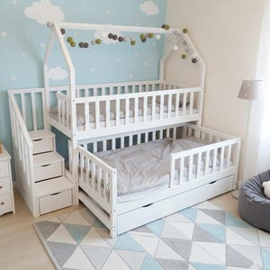 Puede incluir: Una litera de madera blanca con forma de casa, con una escalera con cajones. La cama tiene dos niveles con ropa de cama gris. La habitación tiene una pared azul claro con calcomanías de nubes y estrellas, y una alfombra con estampado geométrico.