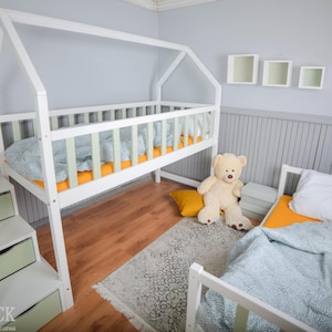 Puede incluir: Una cama alta de madera blanca con estructura en forma de casa, una cama inferior y una escalera con cajones. Las camas tienen ropa de cama azul y amarilla. Un oso de peluche y una pequeña alfombra están en el suelo de madera.