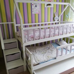 Puede incluir: Una litera blanca con estructura en forma de casa, con una escalera con cajones. La cama tiene dos niveles con ropa de cama y una cama nido extraíble. El fondo tiene un papel pintado a rayas en verde, morado y blanco.