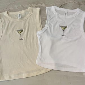 Puede incluir: Dos camisetas sin mangas recortadas, una en crema y otra en blanco, ambas con un diseño de copa de martini bordado.