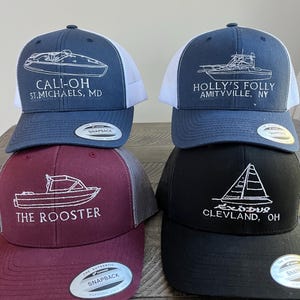 Op de afbeelding: Vier verschillende geborduurde baseballpetten met boot- en zeilbootontwerpen. De petten zijn marineblauw, zwart, kastanjebruin en wit met mesh achterpanelen. De tekst op de petten luidt "Cali-Oh St. Michaels, MD", "Holly's Folly Amityville, NY", "The Rooster" en "Excalibur Cleveland, OH".