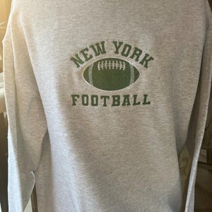 May include: Gray sweatshirt with a green embroidered design that says "NEW YORK FOOTBALL" with a football graphic.