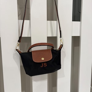 May include: A small black nylon handbag featuring brown leather trim, a top handle, and a detachable shoulder strap. The front flap has a gold-tone snap button closure, and the letters JS are embroidered in brown thread on the black nylon body.