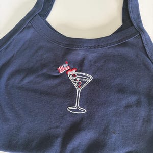 May include: A navy blue tank top featuring an embroidered graphic of a martini glass with red olives and a small American flag toothpick. The design is centered on the chest area of the garment, which has wide shoulder straps and a round neckline.