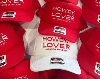 May include: A collection of red and white trucker hats featuring mesh backs and snap closures. Each hat displays the embroidered text "HOWDY, LOVER" followed by "JACKSON HOLE, WY" on the front foam panel. Several hats include an Otto brand label.