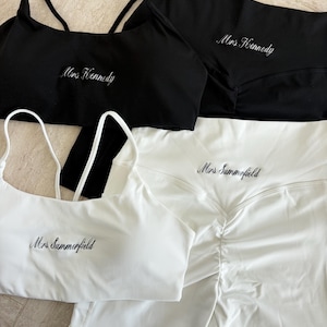 May include: A black sports bra and matching shorts set embroidered with "Mrs. Kennedy" in white, alongside a white sports bra and matching shorts set embroidered with "Mrs. Summerfield" in black. Both sets feature thin straps and ruched detailing.