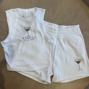 Op de afbeelding: Witte tweedelige lounge set met een tanktop en shorts. De tanktop heeft de tekst "A Bride in a tini bit" en een martini glas afbeelding. De shorts hebben ook een martini glas afbeelding.
