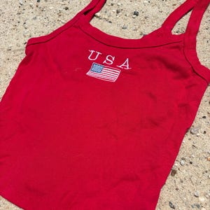 May include: A bright red sleeveless tank top featuring a scoop neckline and thick shoulder straps. Centered on the chest is an embroidered design consisting of the white text USA positioned directly above a small, rectangular American flag patch.