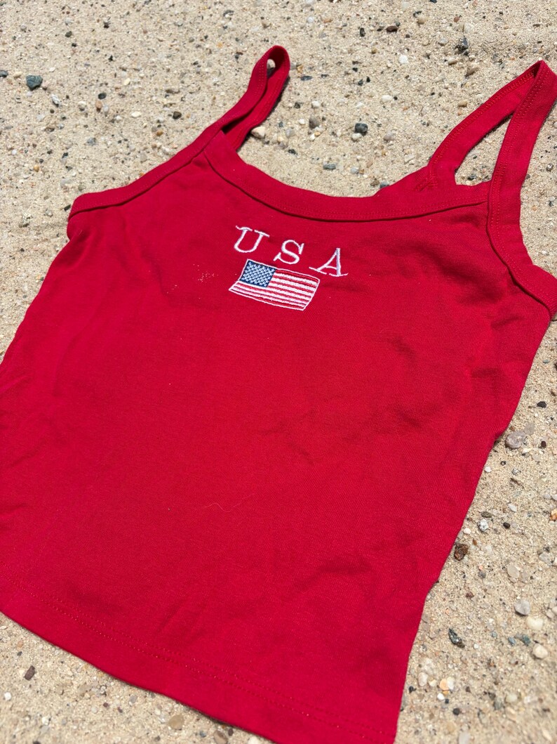 May include: A bright red sleeveless tank top featuring a scoop neckline and thick shoulder straps. Centered on the chest is an embroidered design consisting of the white text USA positioned directly above a small, rectangular American flag patch.