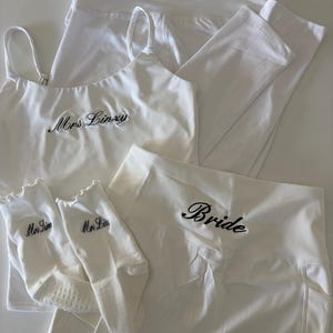 Könnte beinhalten: Ein weißes Set bestehend aus einem Camisole, zwei Leggings und einem Paar Socken. Das Camisole und eine Leggings sind mit dem gestickten Text Mrs Linzy versehen. Die andere Leggings trägt den Text Bride und die Socken zeigen Mrs Linzy.