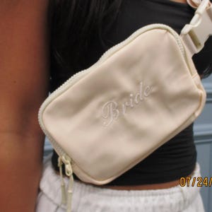 May include: A light beige fanny pack with the word "Bride" embroidered in white script. The bag has a zipper closure and an adjustable strap with a white buckle. The bag is worn across the body.