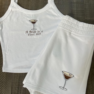Op de afbeelding: Witte tweedelige pyjamaset. De tanktop heeft spaghettibandjes en de shorts hebben een elastische tailleband. Beide stukken hebben een geborduurd martini glas ontwerp. De tanktop heeft de tekst "A Bride in a tini bit".