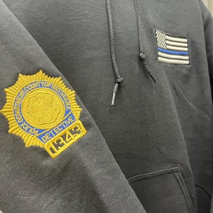 May include: Black hoodie with a gold embroidered police badge on the left sleeve and a thin blue line American flag on the right side. The badge reads "Police Department County of Nassau Detective 1349".