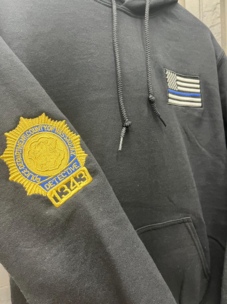 May include: Black hoodie with a gold embroidered police badge on the left sleeve and a thin blue line American flag on the right side. The badge reads "Police Department County of Nassau Detective 1349".