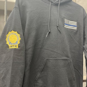 May include: Black hooded sweatshirt with a blue and white American flag patch on the front and a gold embroidered badge on the left sleeve that says "DETECTIVE 18248".