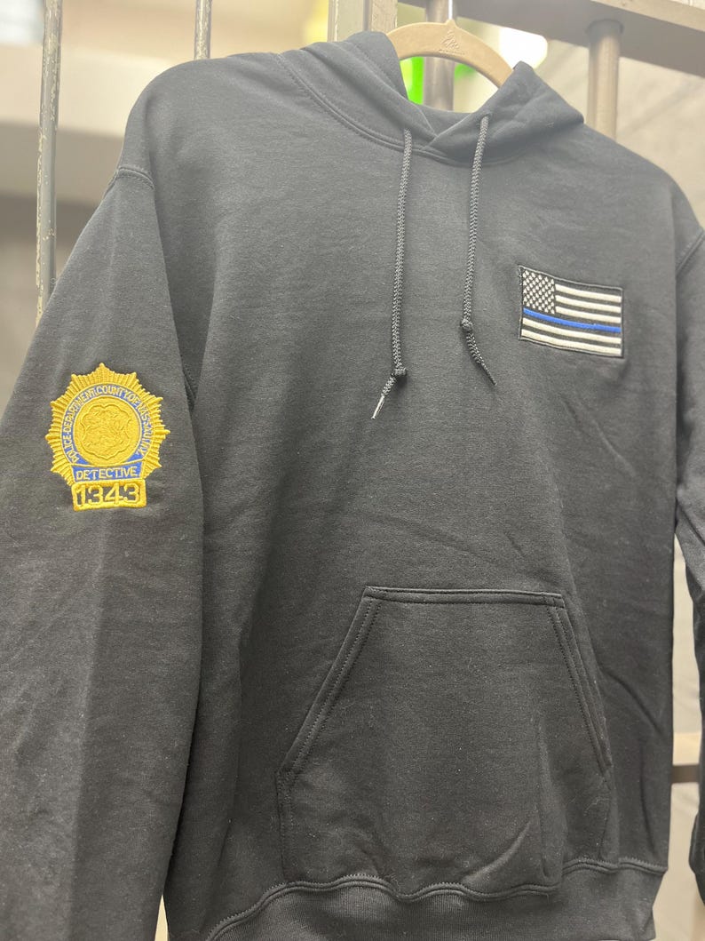 May include: Black hooded sweatshirt with a blue and white American flag patch on the front and a gold embroidered badge on the left sleeve that says "DETECTIVE 18248".