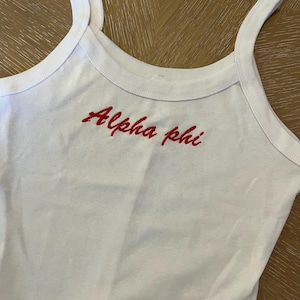 Könnte beinhalten: Weißes Tanktop mit rotem Sticktext "Alpha Phi".