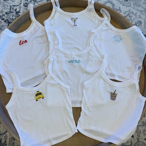 Könnte beinhalten: Fünf weiße Tanktops mit verschiedenen Stickereien. Die Designs beinhalten ein Martini-Glas, eine Muschel, das Wort "mrs", ein Taxi und eine Tasse Eiskaffee.