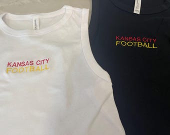May include: A white tank top and a black tank top, each featuring the embroidered text KANSAS CITY FOOTBALL in red and yellow thread. Both garments have a round neckline and a white brand label attached to the inside of the collar area.