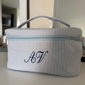 May include: A rectangular cosmetic bag featuring a light blue and white pinstripe pattern. The bag has a top handle, a light blue zipper closure, and the letters AV embroidered in dark blue script on the front center of the exterior fabric.