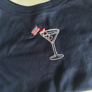 May include: A dark navy blue cotton tank top featuring an embroidered design of a martini glass with three olives and a small American flag toothpick. The embroidery uses white, red, and blue thread to detail the glass, the olives, and the flag.