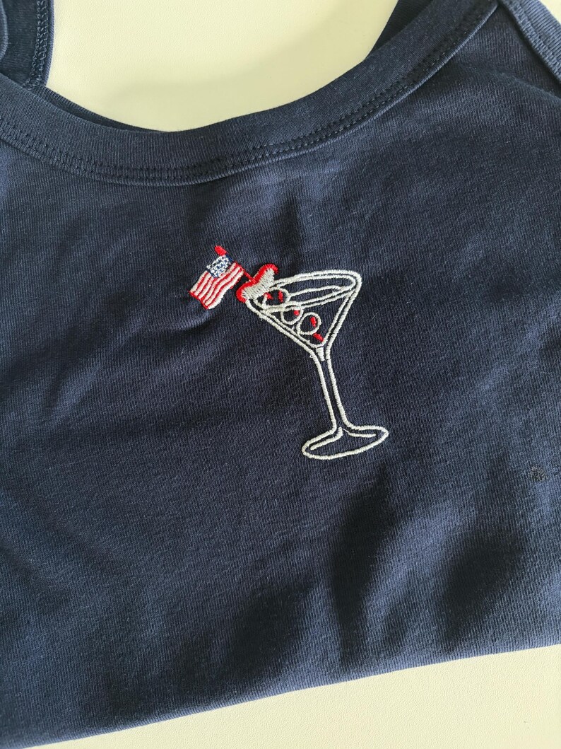 May include: A dark navy blue cotton tank top featuring an embroidered design of a martini glass with three olives and a small American flag toothpick. The embroidery uses white, red, and blue thread to detail the glass, the olives, and the flag.