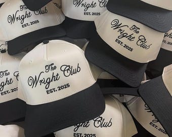 May include: A collection of white and black baseball caps. Each cap features the text "The Wright Club EST. 2025" in black script across the front. The caps have a white front panel and a black brim.