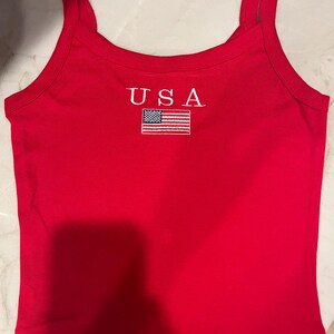 May include: A bright red sleeveless tank top featuring the text USA embroidered in white above a small, rectangular embroidered patch of the American flag. The garment has a scoop neckline and thick shoulder straps, with a clean, simple design.