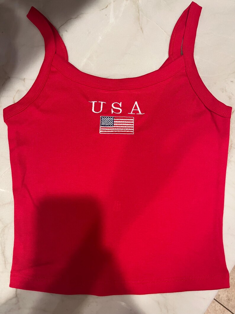 May include: A bright red sleeveless tank top featuring the text USA embroidered in white above a small, rectangular embroidered patch of the American flag. The garment has a scoop neckline and thick shoulder straps, with a clean, simple design.