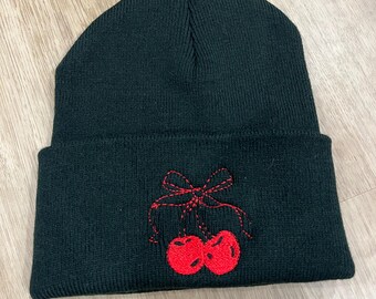 May include: Black beanie hat with a red embroidered cherry design. The hat is made of a soft knit material and has a folded cuff. The cherry design features two red cherries and a red bow.
