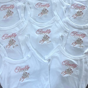 Pode incluir: Uma coleção de regatas brancas caneladas com um design bordado de um buquê de flores com uma fita rosa. O texto "Freshly Picked" é costurado acima das flores em linha rosa, com algumas camisas incluindo texto adicional como "Big", "Little" ou "G-Big".