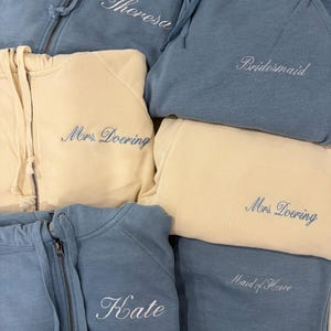 May include: A collection of blue and cream zip-up hoodies and sweatpants featuring embroidered text. The blue garments display the names Theresa, Kate, Bridesmaid, and Maid of Honor, while the cream garments are embroidered with Mrs. Doering.