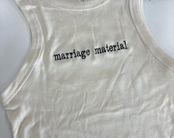 Op de afbeelding: Een crèmekleurige mouwloze tanktop met een geribbelde ronde hals. De voorkant van het kledingstuk is voorzien van de tekst "marriage material" geborduurd in zwarte draad in een typemachine-stijl lettertype. Een wit label is zichtbaar in de nek.