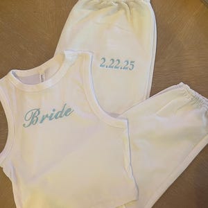 Może przedstawiać: Biały crop top z haftem "Bride" w jasnoniebieskim kolorze. Białe spodnie dresowe z haftem daty "2.22.25" w jasnoniebieskim kolorze.