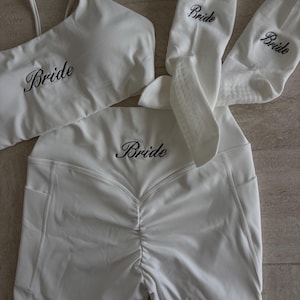 May include: A white athletic set consisting of a sports bra, high-waisted shorts with a ruched back detail, and a pair of socks. Each piece features the word Bride embroidered in black script lettering across the front of the garments.