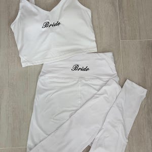 Puede incluir: Un conjunto deportivo blanco de dos piezas que consta de una camiseta sin mangas corta y leggings de cintura alta. La camiseta tiene un escote en V y la palabra Bride bordada en letra negra. Los leggings tienen la palabra Bride bordada en negro.