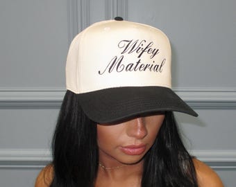 May include: A cream-colored baseball cap with a black brim. The cap features the cursive text "Wifey Material" in black. The hat is worn by a person with long black hair.