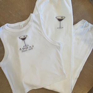 Op de afbeelding: Een wit tanktop en bijpassende joggingbroek met een cocktailglas afbeelding en de tekst "A bride in a tiny bit".