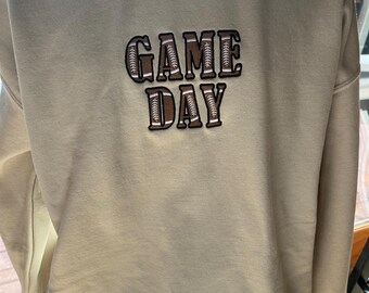 Può includere: Una felpa beige con le parole "GAME DAY" ricamate sul davanti in lettere marroni con cuciture bianche. Le lettere sono contornate da un motivo a pallone da calcio marrone.