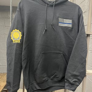 May include: Black hooded sweatshirt with a blue and white American flag patch on the front and a gold embroidered badge on the left sleeve. The badge reads "DETECTIVE 12243".