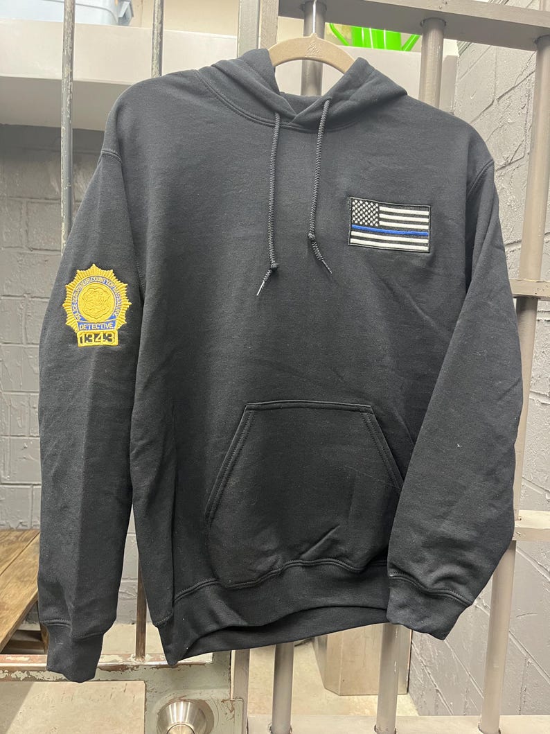 May include: Black hooded sweatshirt with a blue and white American flag patch on the front and a gold embroidered badge on the left sleeve. The badge reads "DETECTIVE 12243".
