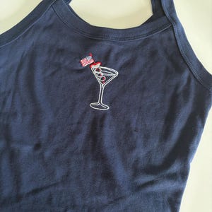 May include: A navy blue halter top featuring an embroidered graphic of a martini glass with three olives on a toothpick. A small American flag is attached to the toothpick. The garment has a round neckline and thick straps.