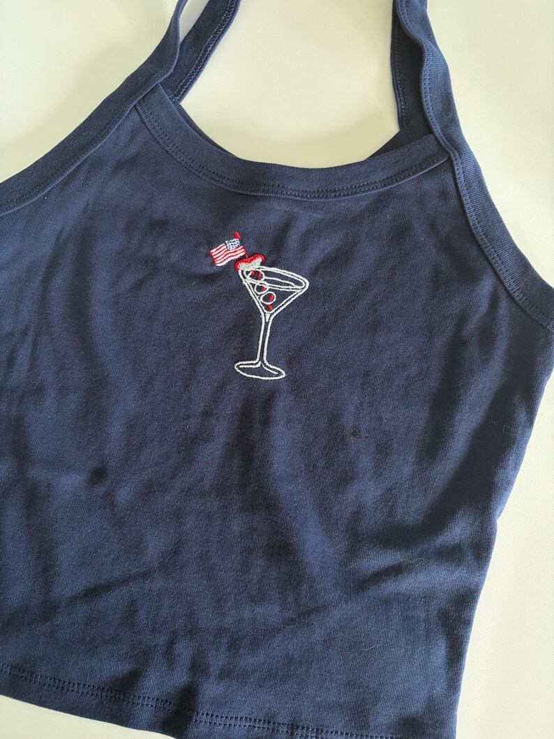 May include: A navy blue halter top featuring an embroidered graphic of a martini glass with three olives on a toothpick. A small American flag is attached to the toothpick. The garment has a round neckline and thick straps.