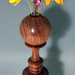 May include: A turned wooden vase with a spherical top section and a wide, tiered base. The wood features a rich brown grain pattern. A small bouquet of flowers is arranged inside, including a large orange daisy, yellow petals, and purple stems.