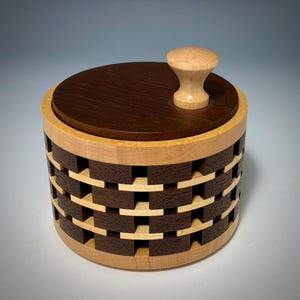 May include: A cylindrical wooden container featuring a woven-style lattice pattern created with alternating light and dark wood blocks. The container has a flat, dark circular lid topped with a small, light-colored wooden knob handle.
